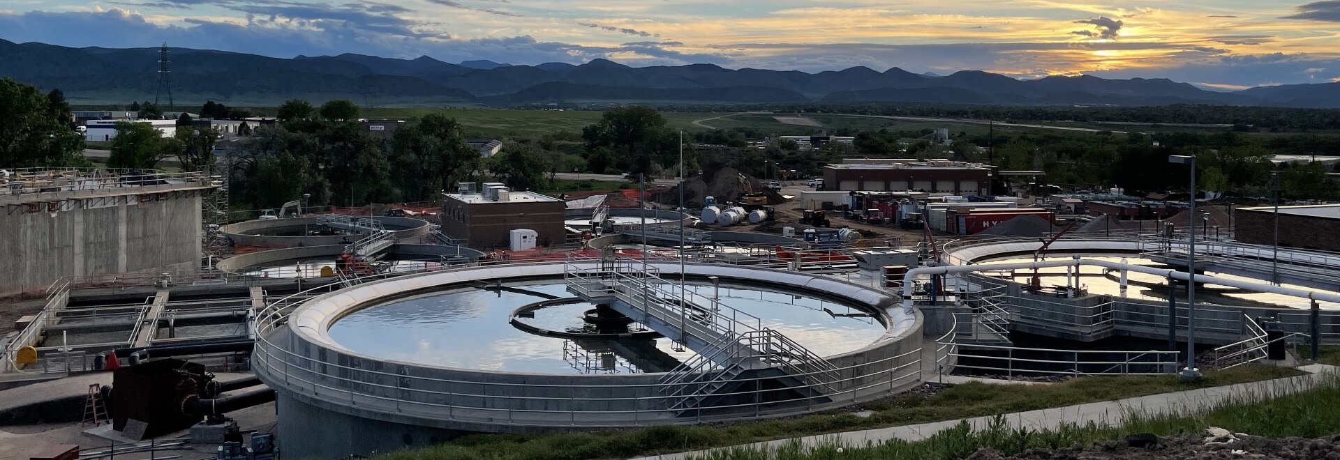 Wastewater treatment plant clarifiers in the foreground surrounded by other buildings at the plant. Scenic mountains and sunset in the background.