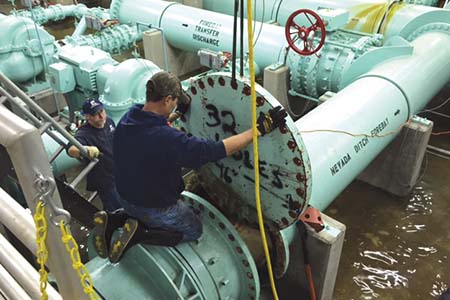 Two staff working, one sitting on his knees on top of a large green pipe holding a disc he is placing 