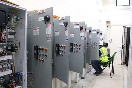 Back of a staff member in a safety vest and hard hat, working in front of a row of electrical equipment