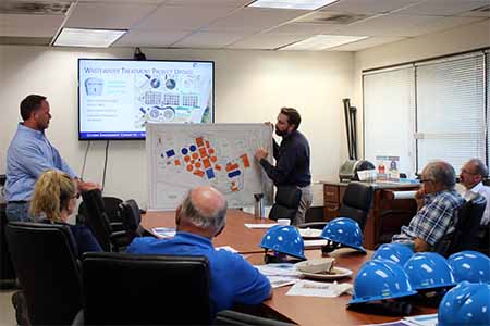 Man pointing out items on a large map to a group of people sitting around a table